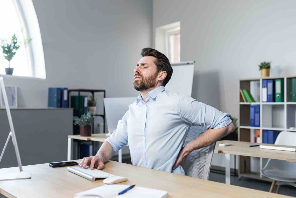 Hombre en oficina sufriendo de dolor de espalda baja, lo que resalta la necesidad de prevención y correcta ergonomía en el lugar de trabajo